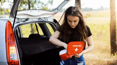 Young woman stands by an open car trunk in a park, focused on a red first aid kit. Sunlit trees in the background convey a calm, prepared mood.